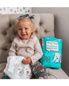 A little girl sits on a chair beside a colorful Mum & You nappy bag, smiling and enjoying her surroundings.