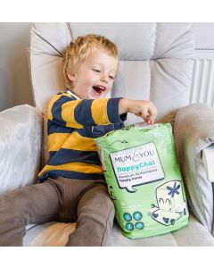 A young boy seated in a chair next to a colorful Mum & You nappy bag, looking happy and relaxed, enjoying a playful moment.