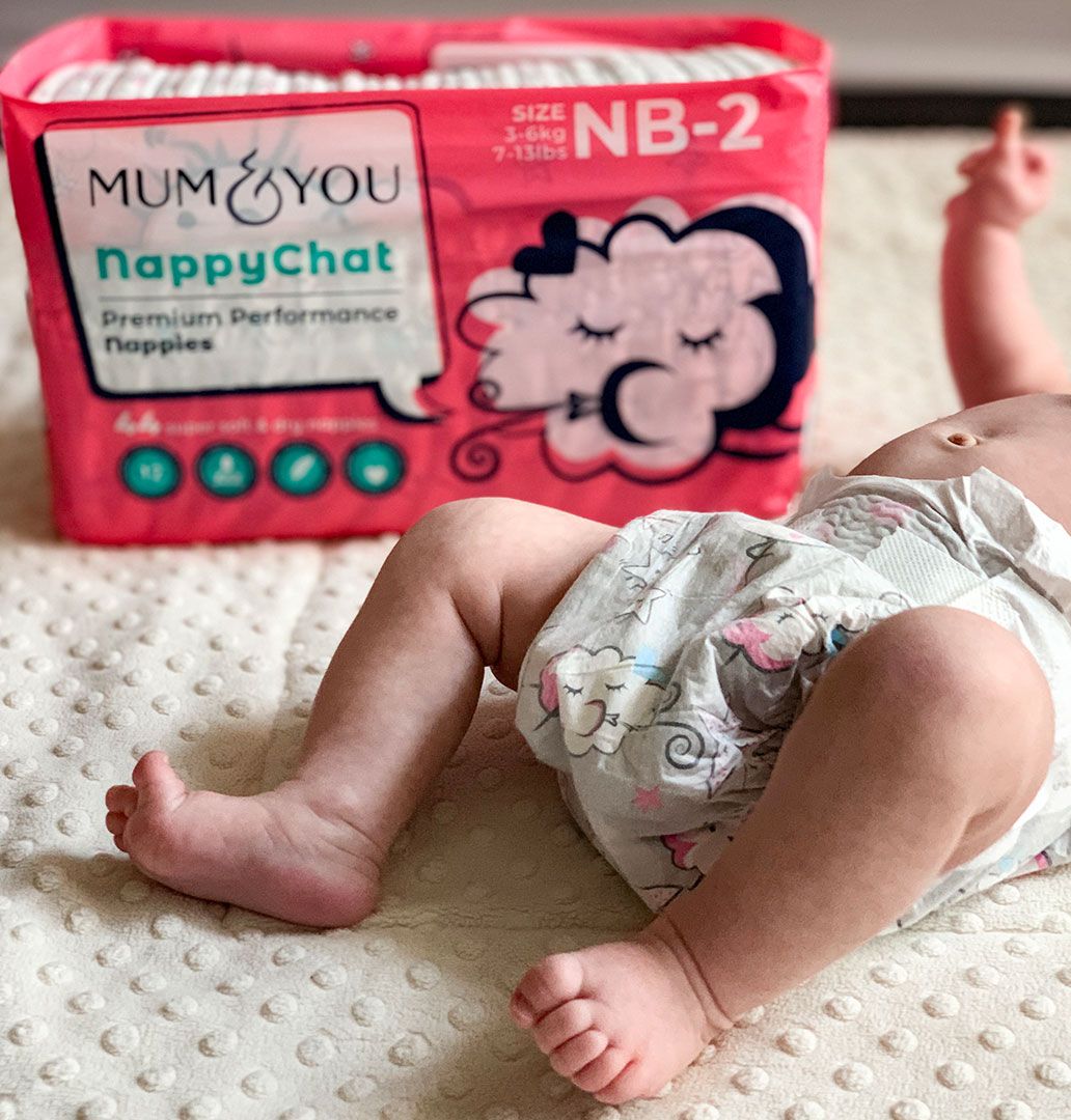 A baby lies on a bed in Mum & You Eco Nappies next to a box of colorful Nappychat designs—soft, breathable, and perfect for playful bonding!